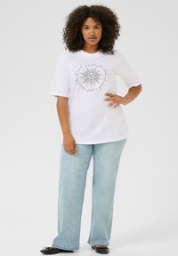T-shirt en coton blanc à manches courtes avec un graphique de soleil noir et du texte, associé à un jean large bleu clair et des ballerines noires.
