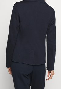 Donna che indossa un blazer fitted blu scuro e pantaloni abbinati, vista da dietro su uno sfondo chiaro e semplice.