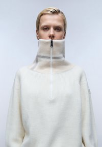 Witte pullover met hoge hals, geribbelde textuur, halve rits en subtiele beige patroonaccenten op de kraag en schouders.