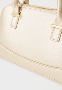 Sac à main beige en matériau synthétique texturé. Doté de quincaillerie dorée, d'une forme structurée, de deux poignées et d'une fermeture à glissière.