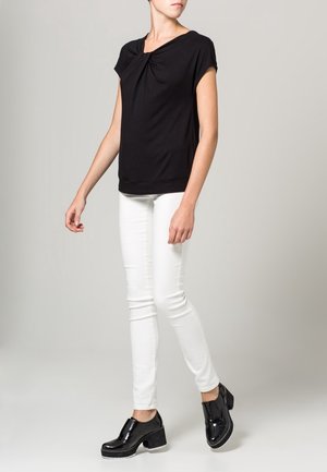 Nainen, joka käyttää mustaa, lyhythihaista yläosaa, jossa on kerätty pääntie, valkoiset kapealahkeiset housut ja mustat korolliset loaferit, seisoo yksiväristä taustaa vasten.