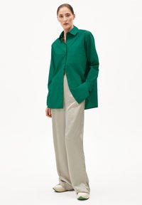 Grünes Button-Up-Hemd aus weichem Material mit einer einzelnen Brusttasche. Getragen mit hellbeige weit geschnittenen Hosen und weißen Schuhen mit grünen Akzenten.