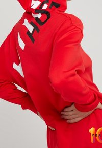 Persoon in een rode hoodie met zwarte en witte tekst, handen op de onderrug geplaatst, zijaanzicht tegen een effen achtergrond.