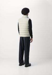 Light-colored puffer vest with a high collar, featuring horizontal quilted sections; worn over a black long-sleeve shirt and striped trousers.