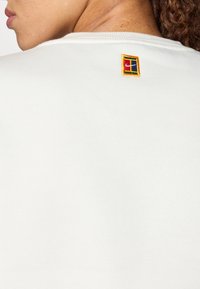 Λευκό φούτερ με ριγωτό λαιμό, που διαθέτει μια μικρή πολύχρωμη κεντημένη ετικέτα στο πίσω μέρος κοντά στον γιακά. Μαλακή υφή.