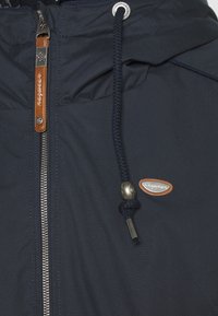 Marineblå jakke lavet af nylon, med en sølvfarvet lynlås, brune læderdetaljer, justerbar snoretræk og et logo-patch.