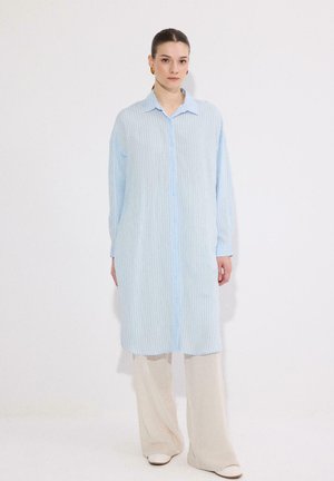 Nő világoskék, hosszú, csíkos ingruha, bézs bőszárú nadrág és fehér cipő viselése közben, egyszínű fehér háttér előtt áll.