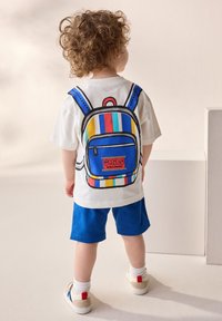 White t-shirt featuring a colorful backpack graphic with blue, yellow, and red stripes, paired with blue shorts and light sneakers.
