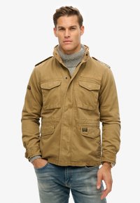 Tan canvas jack met meerdere voorzakken, epauletten op de schouders en een hoge kraag, gedragen over een grijze coltrui, gecombineerd met blauwe jeans.