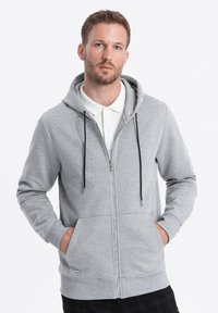 Sweat à capuche gris zippé avec cordons noirs ajustables, poches avant, poignets côtelés et texture douce, porté sur un polo blanc.