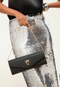 Juodas rankinės krepšys su auksine grandine, tekstūruota paviršiumi ir dekoratyvine leopardine galvos uždanga. Aplink yra sidabrinė sijonas su blizgučiais.
