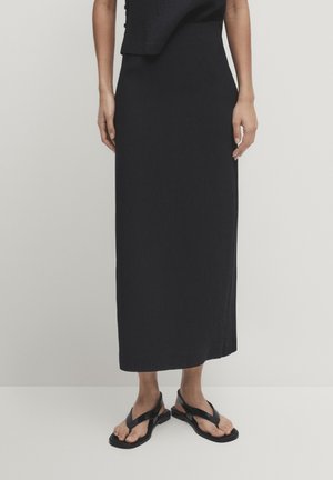Femme portant une jupe midi noire et des sandales fines noires, debout sur un sol blanc, les mains détendues le long du corps.