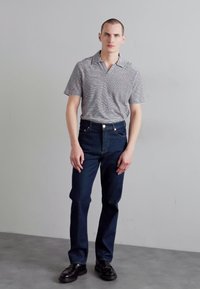 Camisa polo de manga corta a rayas en blanco y negro, combinada con jeans de mezclilla azul oscuro. El modelo lleva zapatos negros y está de pie frente a un fondo gris.