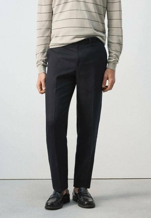 Osoba ubrana w czarne, szyte na miarę spodnie, czarne skórzane mokasyny i beżową koszulę z długim rękawem z cienkimi, poziomymi paskami.