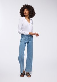 Camicia bianca a bottone, jeans a gamba larga blu chiaro con due tasche frontali e sandali neri con borchie decorative. Tessuto morbido e vestibilità rilassata.