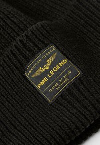 Zwarte gebreide beanie met ribbeltextuur. Voorzien van een rechthoekig stoffen label in zwart en geel met tekst en vleugeldiagrammen.