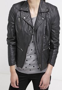 Personne portant une veste de motard en cuir noire par-dessus un pull gris à pois noirs, associée à un pantalon noir, debout devant un fond uni.