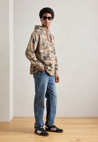 Beige printed hoodie with red drawstrings, paired with light blue jeans and black sneakers featuring white stripes. Smooth texture, casual design.
