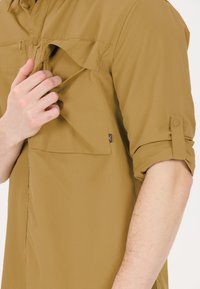 Tan knoopshirt gemaakt van lichtgewicht stof, met een voorzak met klep en opgerolde mouwen met knoopjes. Zachte textuur.