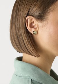 Gold heart-shaped earring with polished surface, featuring a small embossed logo at the center, worn on a short bob hairstyle.