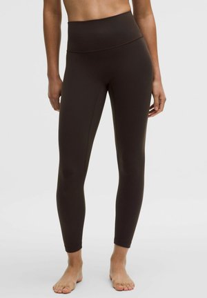 Vrouw die donkerbruine leggings met hoge taille draagt, staat op blote voeten tegen een effen lichtgrijze achtergrond.