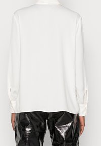 Blusa bianca a maniche lunghe con una texture liscia, caratterizzata da un colletto e maniche con bottoni, abbinata a pantaloni neri lucidi a vita alta.