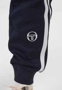 Tamsiai mėlynos sportinės kelnės su baltomis šoninėmis juostelėmis, su apvaliu baltu logotipu. Minkšta tekstūra ir pritaikyta juosta ties kulkšnimis.