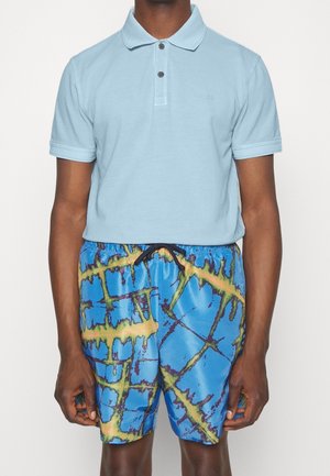 Lys blå polo-shirt med to knapper og et lille logo. Lyseblå badetøj med et rutemønster i grønne og gule nuancer.