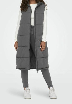 Mujer que lleva un abrigo gris largo acolchado sin mangas sobre una camiseta blanca de manga larga, pantalones grises y zapatillas blancas, de pie con las piernas separadas.