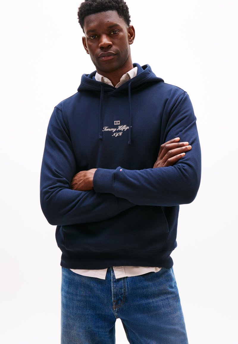 Hoodie in cotone blu navy con coulisse e logo ricamato sul petto, indossato sopra una camicia con colletto, abbinato a jeans in denim blu.