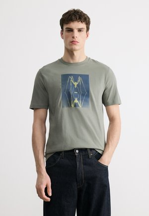 Nuori mies, jolla on päällään vihreä T-paita, jossa on keltainen abstrakti hämähäkkikuvio, ja tummansiniset farkut, seisomassa yksinkertaista vaaleaa taustaa vasten.