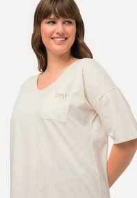 Lys beige oversized t-shirt med v-hals, korte ærmer og en brystlomme med ordet "KÆRLIGHED" broderet i lys beige.