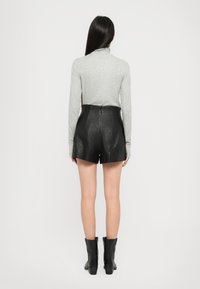 Donna con capelli lunghi e scuri, che indossa un top grigio a maniche lunghe, pantaloni in pelle nera e stivali neri alla caviglia, in piedi di spalle contro uno sfondo bianco.