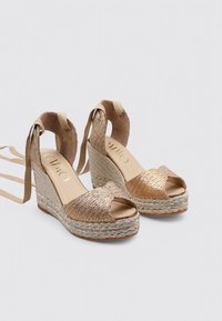 Sandalias de cuña con suela de yute trenzado, diseño de punta abierta y acentos metálicos dorados. Cierres en el tobillo para un ajuste seguro.