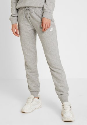 Personne portant un pantalon de survêtement Nike gris clair avec taille à cordon et des baskets blanches sur un fond blanc uni.