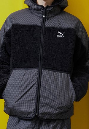 Svart och grå dragkedjetröja med texturerad fleece och tryckt tyg. Har en hög krage och ett varumärkeslogotyp på bröstet.