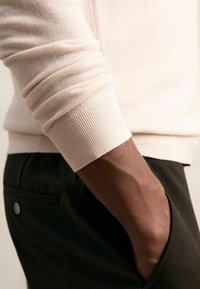 Heller beiger Pullover mit gerippten Bündchen, kombiniert mit dunkler Hose, zeigt eine Nahaufnahme des Arms und der Hand in einer Seiten-Tasche.