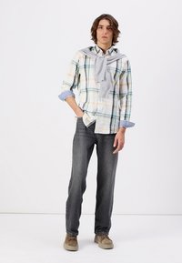 Chemise à carreaux blanche, verte et jaune, associée à un jean gris foncé. Un pull gris clair drapé sur les épaules. Chaussures beiges.