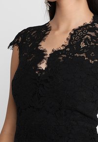 Schwarzes Spitzenkleid mit kurzen Flügelärmeln, das einen gewellten Ausschnitt und filigrane florale Muster zeigt. Der Stoff hat ein strukturiertes, geschichtetes Aussehen.