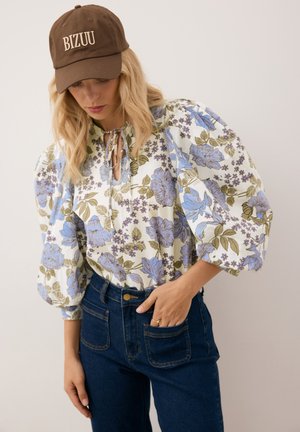 Bloes met bloemenprint en opgeblazen mouwen, met blauwe en groene bloemen op een crèmekleurige achtergrond. Gecombineerd met high-waisted spijkerbroeken en een bruine pet.