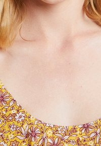 Gul tyg med blommönster som innehåller vita och rosa blommor. Halsringningen har en mjuk scoop-form, och texturen ser slät ut.