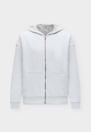Junger Mann trägt einen hellgrauen Kapuzenpullover mit Reißverschluss, weißes T-Shirt, dunkelblaue hochgekrempelte Jeans und weiße Turnschuhe und steht vor weißem Hintergrund.