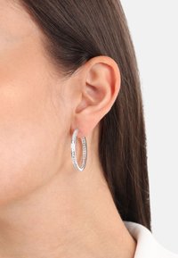 Pendiente de aro de plata con un diseño de doble lazo, adornado con piedras transparentes a lo largo del borde exterior, acabado suave y superficie pulida.