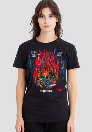 Frau trägt ein schwarzes Stranger Things T-Shirt mit einer flammenden Spinnenklaue, die eine Person hält, und dem Text "Bald ist alles vorbei" und "Die Brücke."