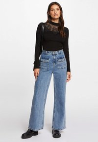 Top negro de manga larga con detalles de encaje, combinado con jeans de pierna ancha de cintura alta en azul claro. Botines gruesos negros. Bolsa negra simple.