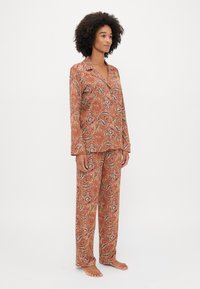 Červený pyžamový set s paisley vzorem, který obsahuje košilový top s knoflíkovým zapínáním a odpovídající kalhoty. Hladká tkanina s tekoucím drapováním a volným střihem.