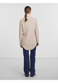 Beżowy prążkowany sweter z wysokim kołnierzem, dłuższym tylnym brzegiem i długimi rękawami, zestawiony z ciemnoniebieskimi szerokimi dżinsami i czarnymi butami.