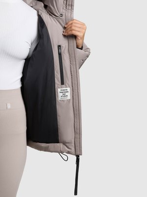 Doudoune, extérieur gris clair, doublure intérieure noire, poche zippée visible, et une étiquette avec la mention « Un câlin chaud fait de tissu. »