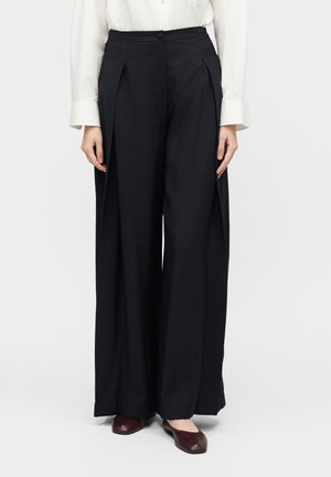 Femme portant un pantalon noir large taille haute associé à un chemisier blanc à manches longues et des chaussures plates marron foncé, debout droite.