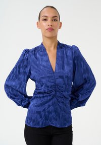 Blue blouse with a deep V-neck, gathered front, and voluminous sleeves. Made from textured fabric with a subtle pattern.